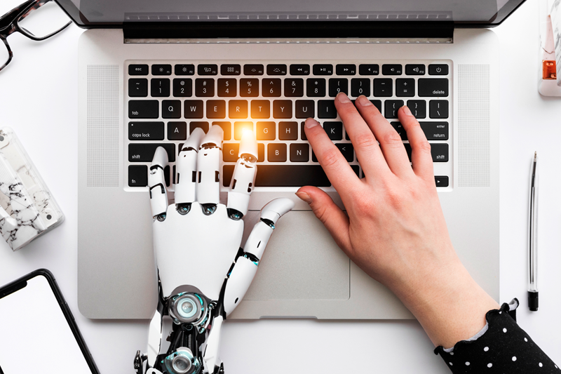 Imagem mostra uma mao robótica e uma mao humana trabalhando juntas em um laptop